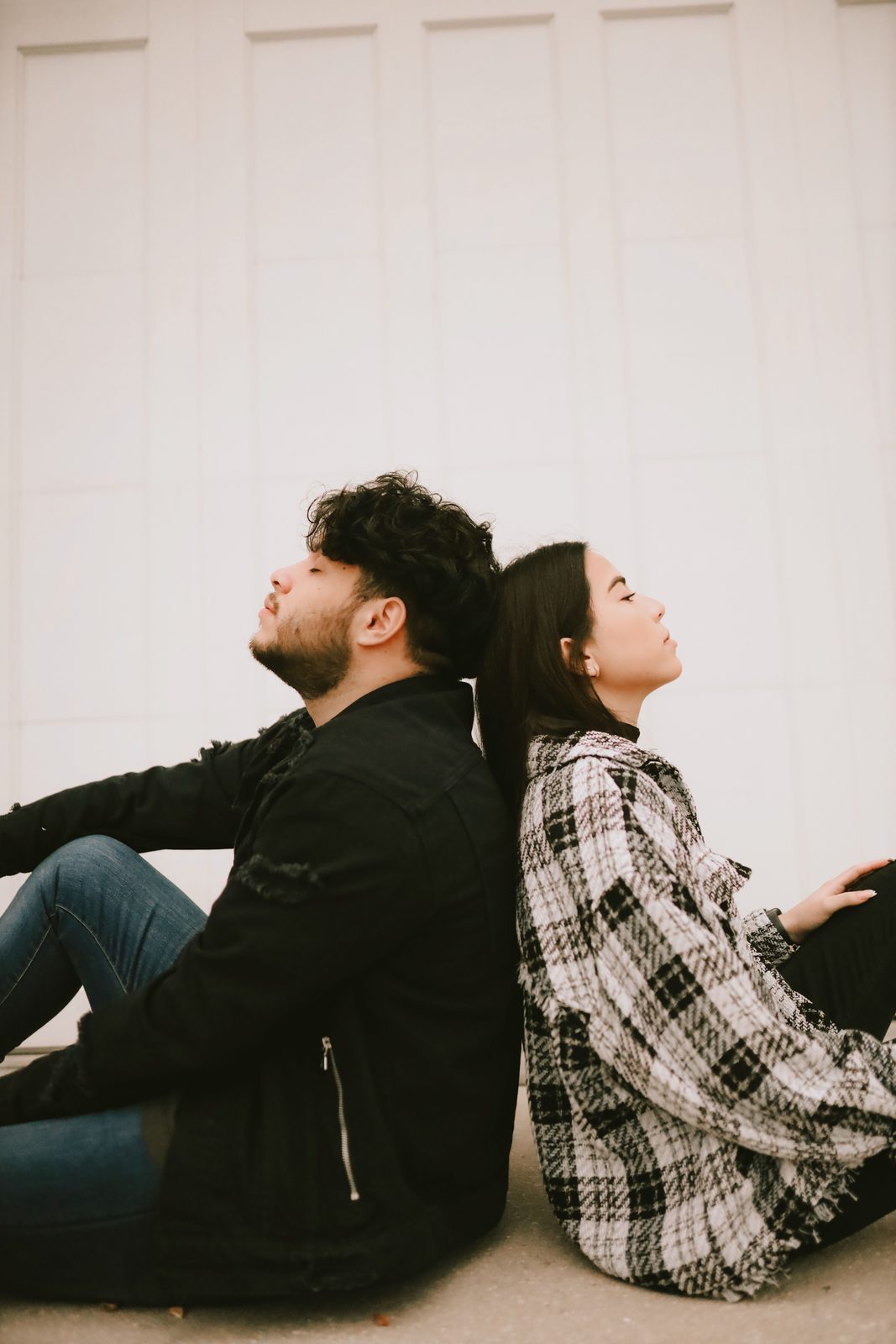 A couple sitting back to back against a pale wall, eyes closed